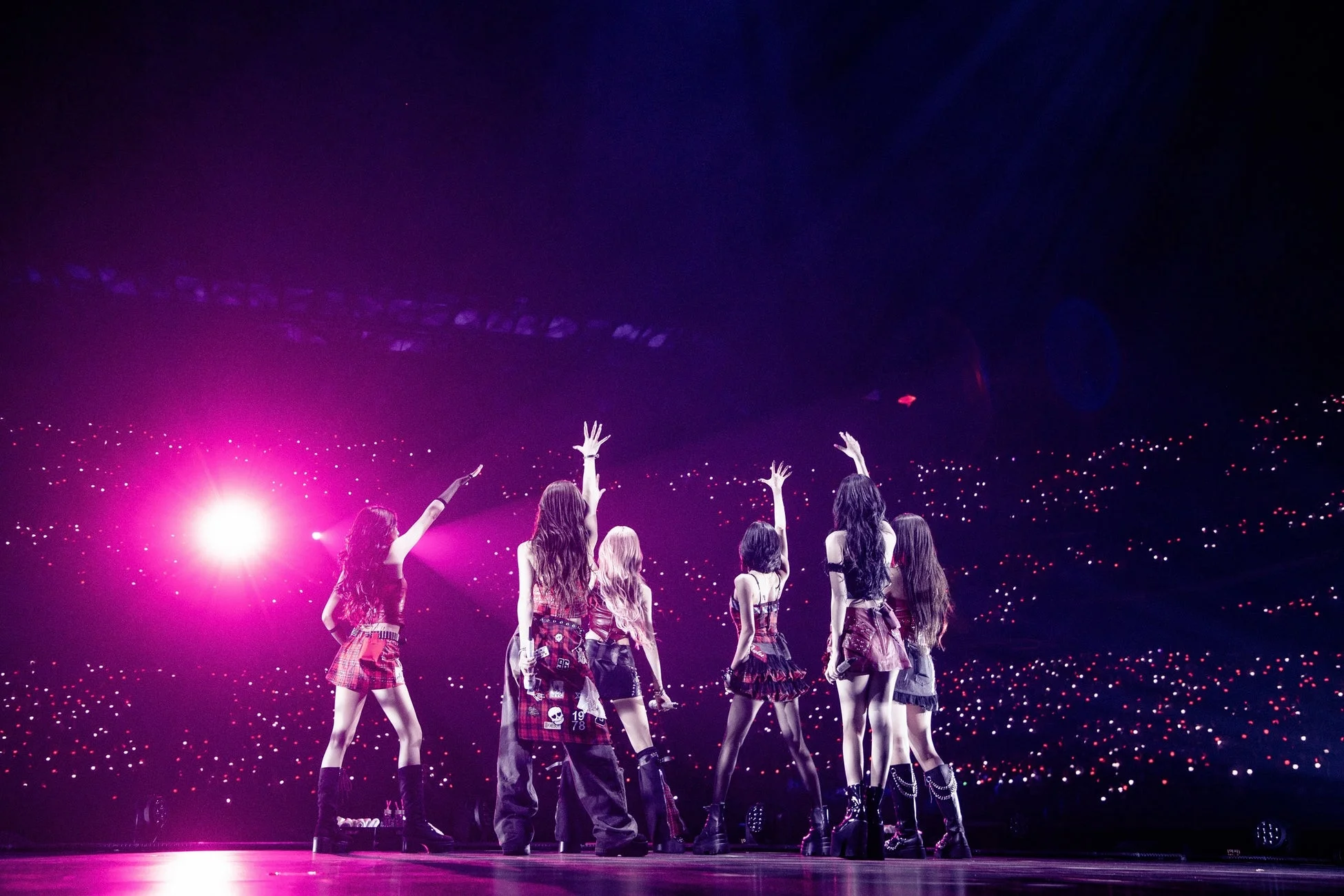 ステージ上でパフォーマンスを行う複数の女性アーティストの後ろ姿が写っています。彼女たちは観客に向かって手を挙げ、会場全体はピンクや紫の照明と、無数の観客の持つライトで幻想的に彩られています。