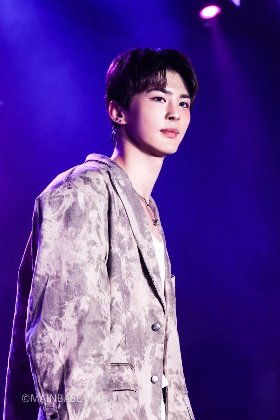 紫色の照明が照らすステージに立つ若い男性のポートレート。タイダイ柄のジャケットを着用し、青い瞳でクールな表情を見せています。