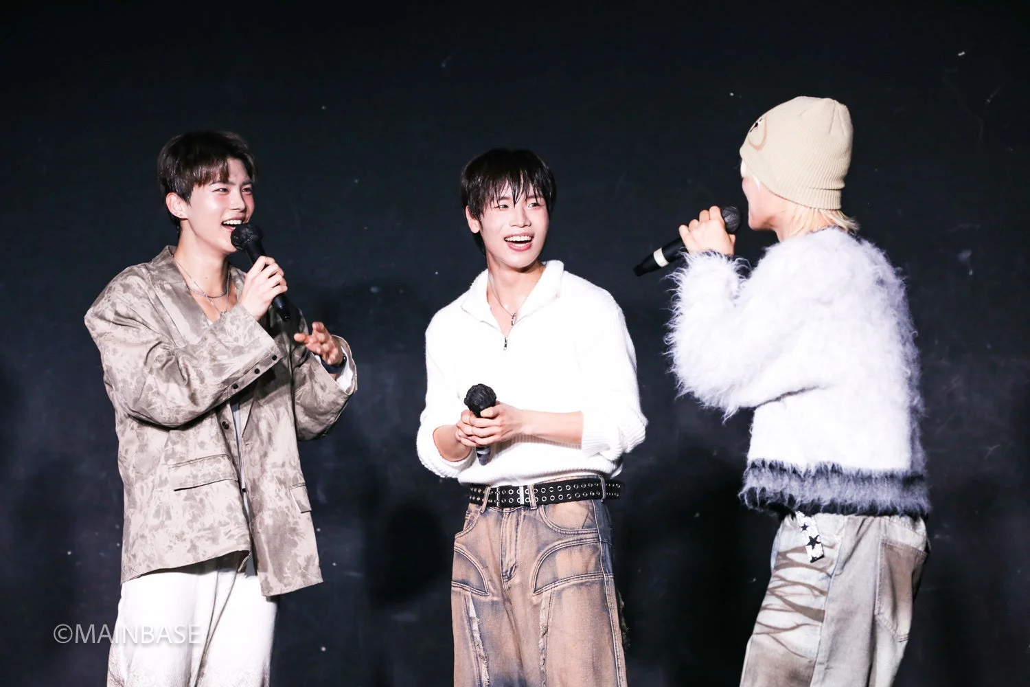 暗い背景のステージで、3人の若者がマイクを持ち、笑顔で話している様子です。彼らは楽しそうに交流しており、音楽イベントやパフォーマンスの一幕を捉えたものと思われます。