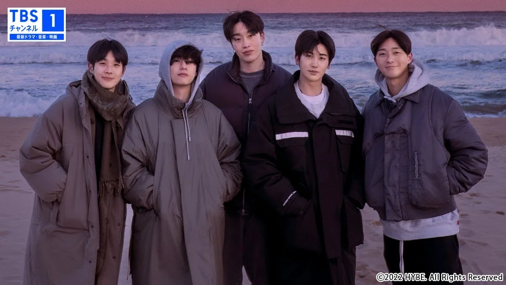 冬の海辺で厚手のコートを着た5人の男性が並んで立っています。背景には波打ち際と夕焼け空が見え、和やかな雰囲気が漂っています。