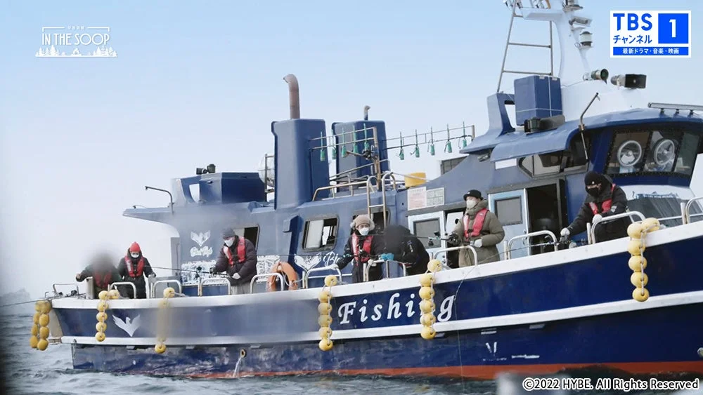 寒い曇り空の下、ライフジャケットとマスクを着用した人々が青い漁船で釣りをしている。船には「Fishing」の文字があり、テレビ番組「IN THE SOOP」とTBSのロゴが表示されている。