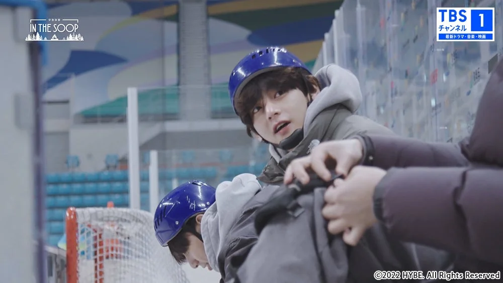 アイススケートリンクでヘルメットを着用した若者たちが、防寒着を着て楽しんでいる様子。一人がもう一人に何か手伝っている瞬間を捉えています。